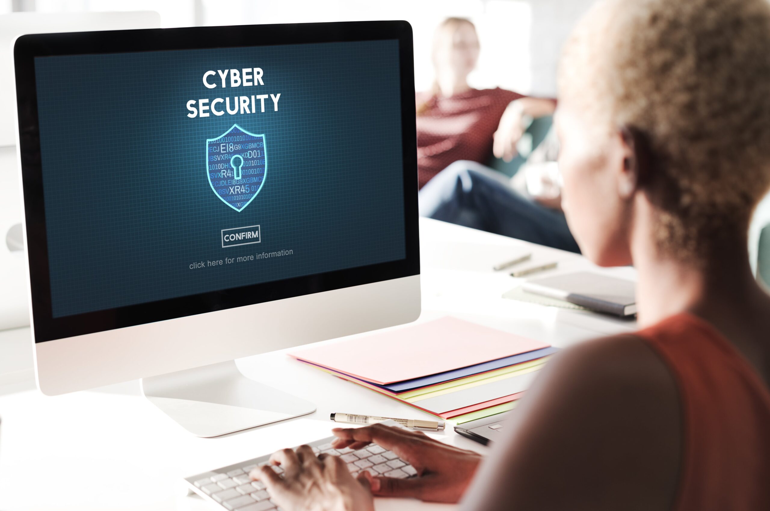 A woman at a desk views a 'Cyber Security' interface on her computer, featuring encryption symbols and a 'CONFIRM' button.